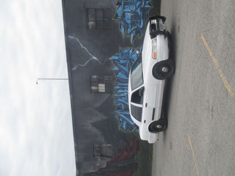 2004 Ford Crown Victoria - A white 2004 Ford Crown Victoria is parked in front of a building with a large mural on its side.