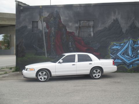 2004 Ford Crown Victoria - A white 2004 Ford Crown Victoria is parked in front of a large mural on a brick wall.