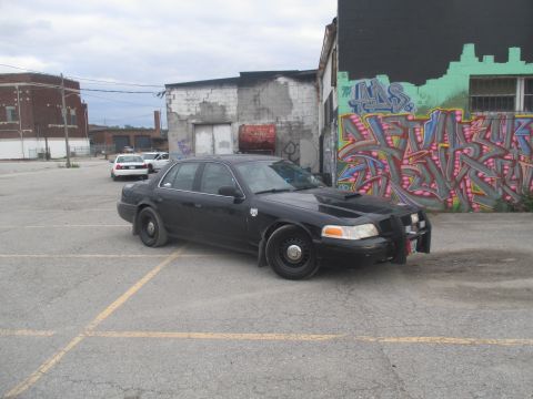 2004 Ford Crown Victoria - A black sedan with a push bar is parked in a lot with graffiti on the wall.
