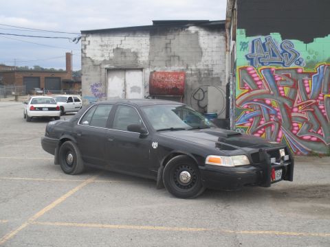 2004 Ford Crown Victoria - A black 2004 Ford Crown Victoria, modified with a push bar and hood scoop, is parked in an urban lot with graffiti on the wall.