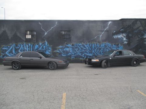 2004 Ford Crown Victoria - Two Ford Crown Victoria police cars are parked in front of a wall with graffiti art.