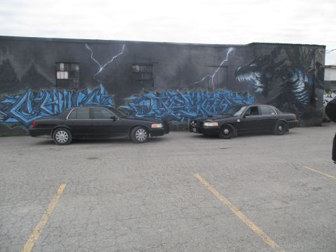 2004 Ford Crown Victoria - Two sedans are parked in front of a building with a large mural of a dragon and graffiti.