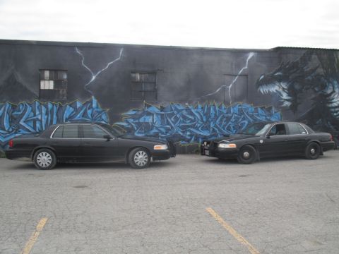 2004 Ford Crown Victoria - Two black Ford Crown Victorias are parked in front of a building with a large mural.