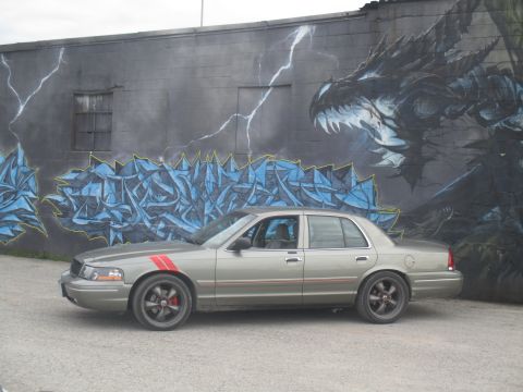 2004 Ford Crown Victoria - A gray Ford Crown Victoria is parked in front of a large mural a dragon and abstract graffiti art.