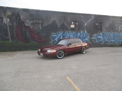 2004 Ford Crown Victoria - A maroon sedan is parked in front of a graffiti-covered wall a large mural.