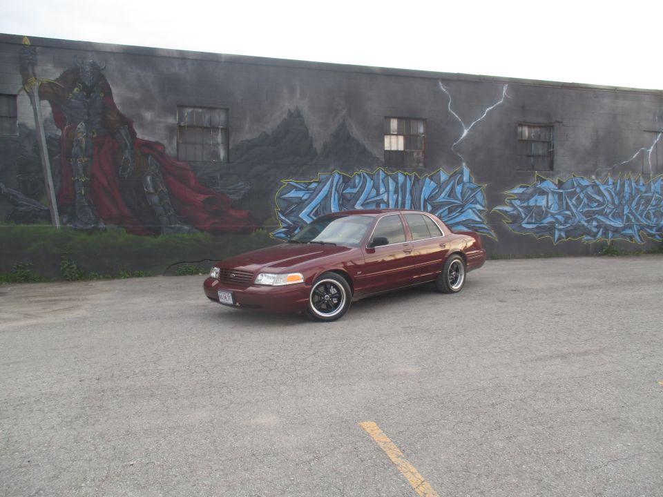 Maroon sedan parked in front of a mural, black rims, custom exhaust tips visible