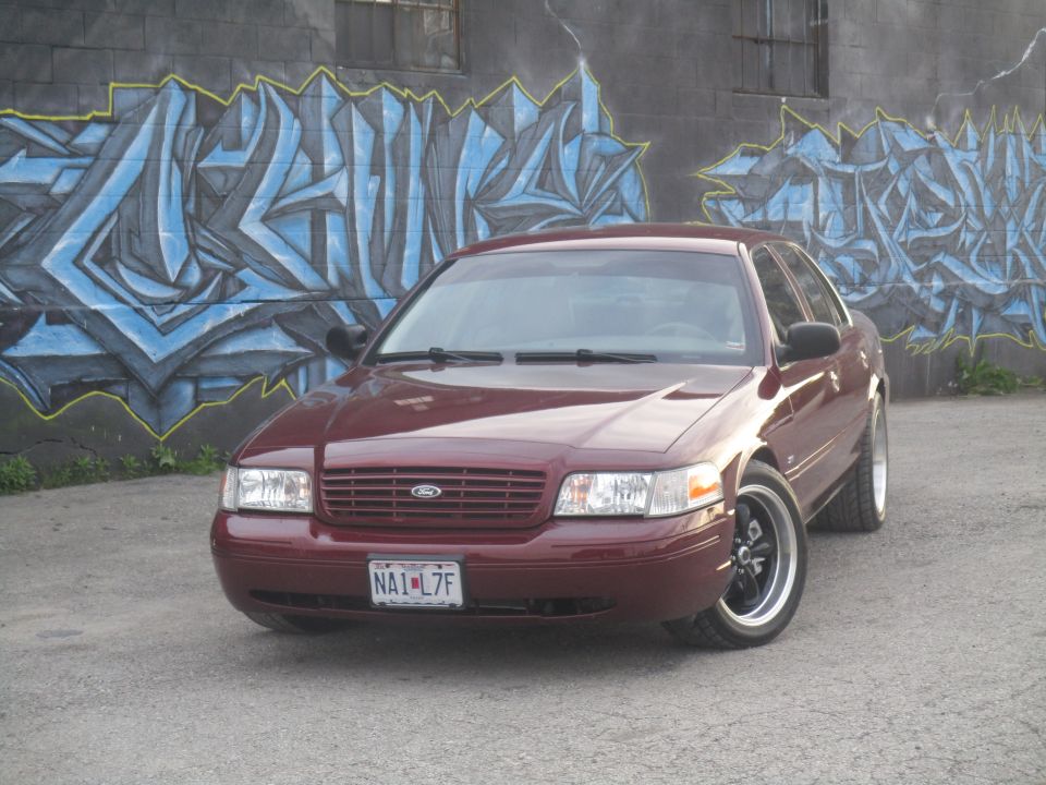 Maroon Ford Crown Victoria parked in front of graffiti wall. License plate NA1-L7F