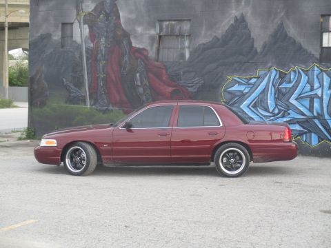 2004 Ford Crown Victoria - A maroon sedan with aftermarket wheels is parked in front of a graffiti-covered wall.