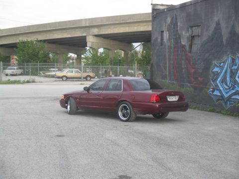 2004 Ford Crown Victoria - A maroon sedan is parked in an urban setting with graffiti on the wall and an overpass overhead.