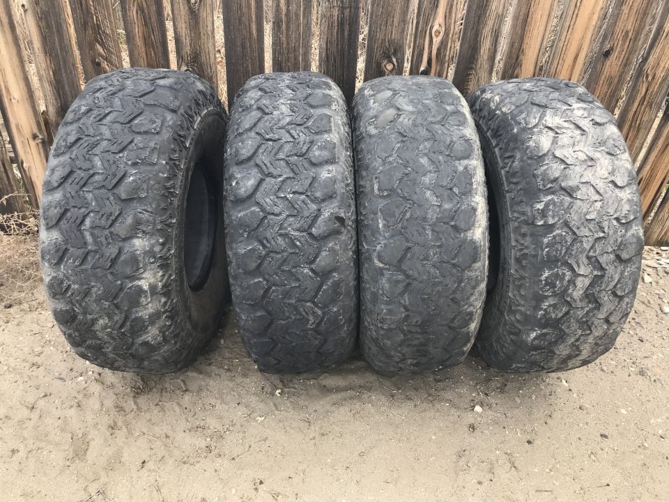 Four used off-road tires leaning against a wooden fence