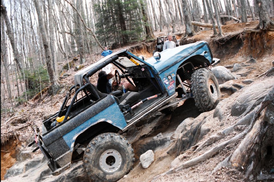 Blue 1976 Ford Bronco off-roading on a rocky incline, with a second vehicle visible behind it