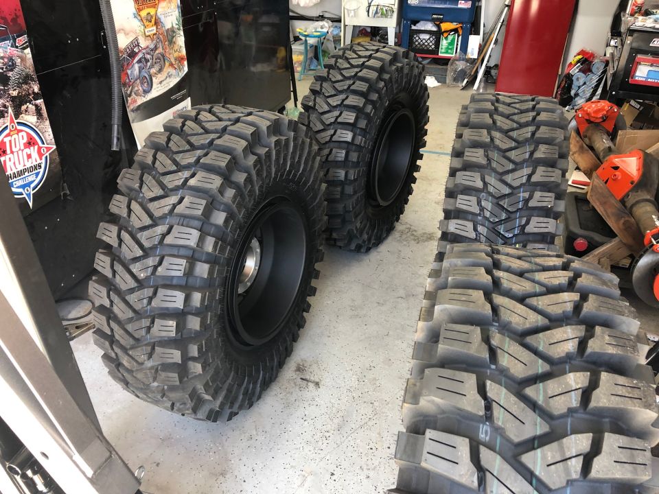 Four large off-road tires with aggressive tread patterns are displayed on a garage floor. One tire is mounted on a black rim