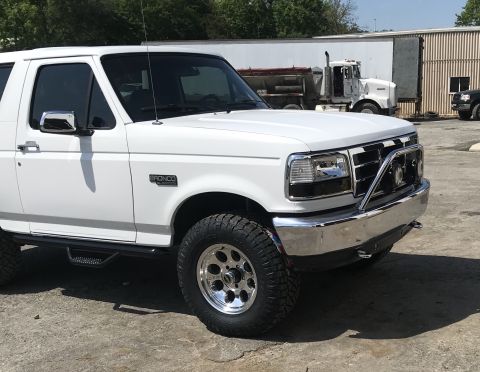 1995 Ford Bronco - A white 1995 Ford Bronco is shown from the front three-quarter view, with other vehicles and buildings in the background.