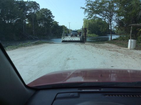 1992 Ford Bronco - A dark SUV is on a ferry, being transported across a waterway surrounded by trees.