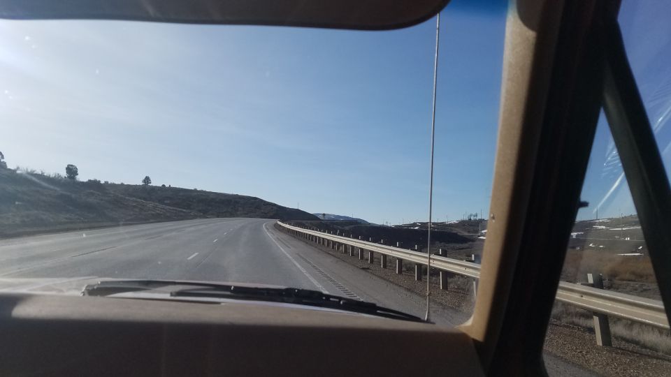 Dashboard and windshield view from inside a vehicle driving on a highway with a guardrail and rolling hills under a clear blue sky
