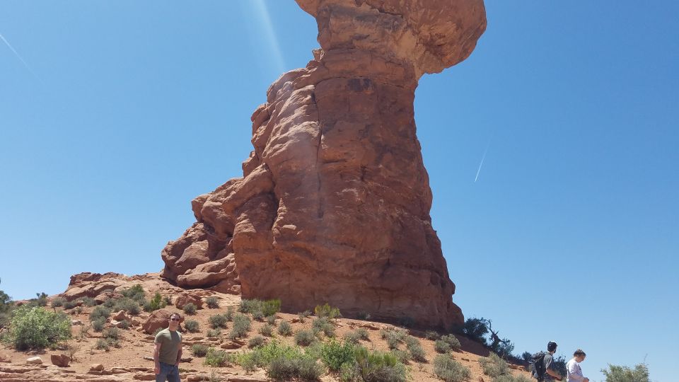 Person stands in desert landscape with large sandstone rock formation under a clear blue sky