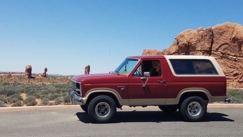 1985 Ford Bronco - This classic Ford Bronco is parked on a road with desert scenery and rock formations under a clear blue sky.