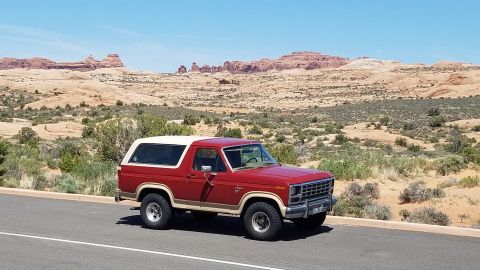 1985 Ford Bronco - Red 1985 Ford Bronco with a white removable top driving through a desert landscape.