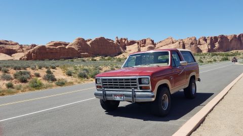1985 Ford Bronco - A red Ford Bronco is parked on the side of a road with a desert landscape and rock formations under a clear blue sky.