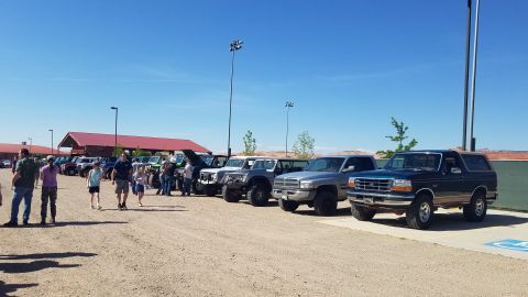 1985 Ford Bronco - A dark blue Ford Bronco is parked in a lot with other vehicles and people under a clear blue sky.