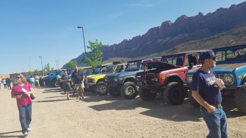 1985 Ford Bronco - A line of classic Ford Broncos are parked in a dusty lot with a scenic mountain range behind them.