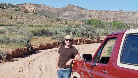 1985 Ford Bronco - A man leans against a red 1980s Ford Bronco in a scenic desert environment.