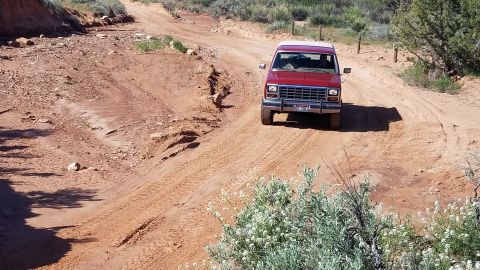 1985 Ford Bronco - A red SUV navigates a dusty, off-road trail with sparse vegetation.