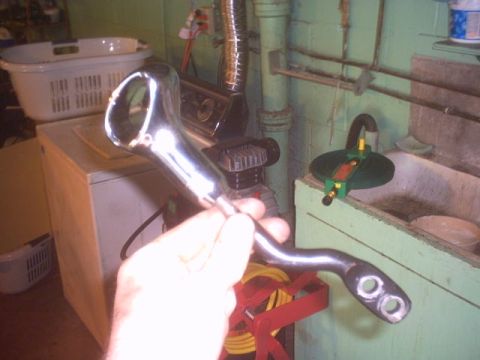 1991 Ford LTD Crown Victoria - A chrome-plated metal part, possibly a linkage or lever, is held in a hand against a background of a laundry basket and plumbing.