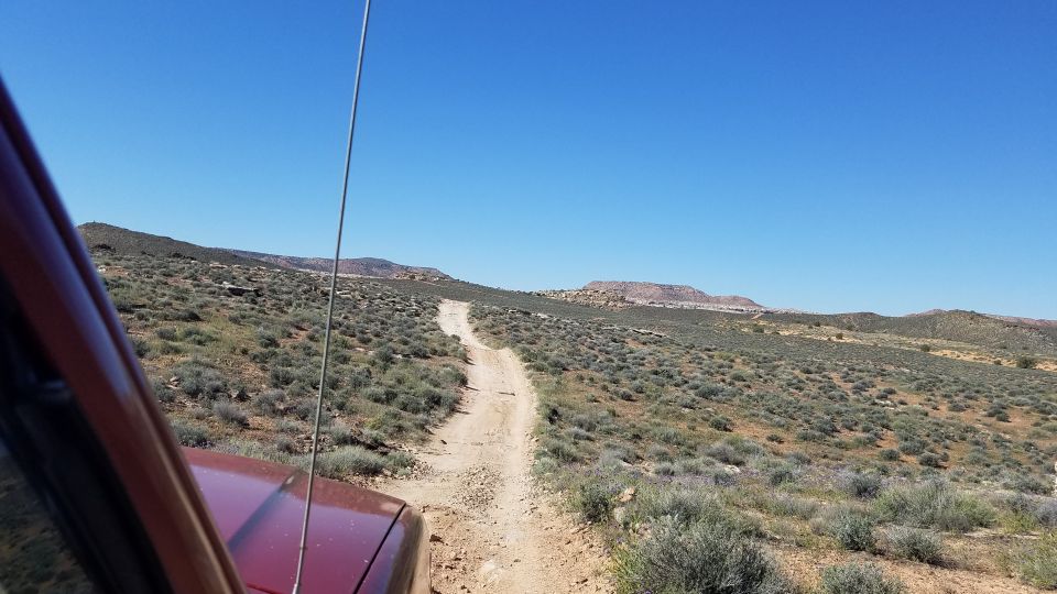 Red SUV's hood and fender visible, driving on a winding dirt road through a desert with scrub brush and distant hills under a clear blue sky