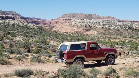1985 Ford Bronco - 1985 Ford Bronco parked on a dirt trail in a desert canyon with red rock formations.