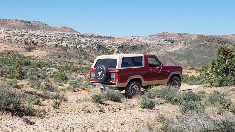 1985 Ford Bronco - A maroon and tan Ford Bronco is parked in a desert landscape with rocky hills in the background.