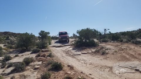 1985 Ford Bronco - This red SUV is navigating a challenging off-road trail with sparse vegetation and rocky ground.
