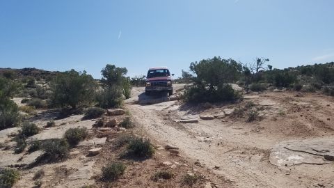 1985 Ford Bronco - A red SUV navigates a challenging off-road path in a desert landscape under a bright blue sky.
