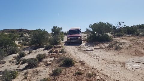 1985 Ford Bronco - A red van navigates a rocky dirt path through a desert terrain under a clear blue sky.