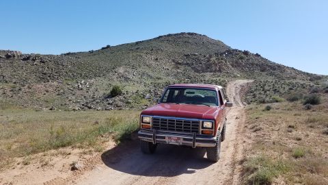 1985 Ford Bronco - 1985 Ford Bronco navigates a remote dirt track through a dry, hilly terrain under a clear blue sky.