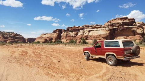1985 Ford Bronco - A red 1985 Ford Bronco sits parked in a desert landscape with sandstone cliffs under a bright blue sky.