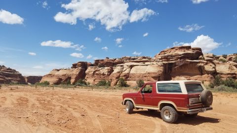 1985 Ford Bronco - A red 1985 Ford Bronco is parked in a desert landscape with red rock formations in the background under a blue sky with white clouds.