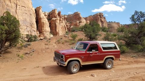 1985 Ford Bronco - A red and tan 1985 Ford Bronco is parked on a dirt road in a desert landscape with large sandstone cliffs in the background.