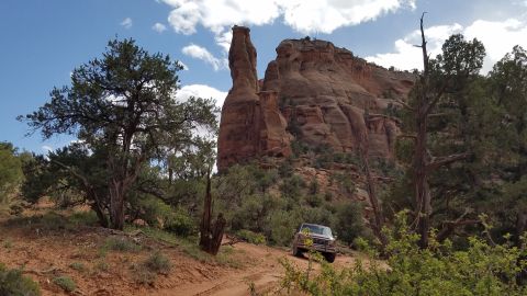 1985 Ford Bronco - A maroon 1985 Ford Bronco is parked on a dirt road in a desert landscape with large rock formations and trees.