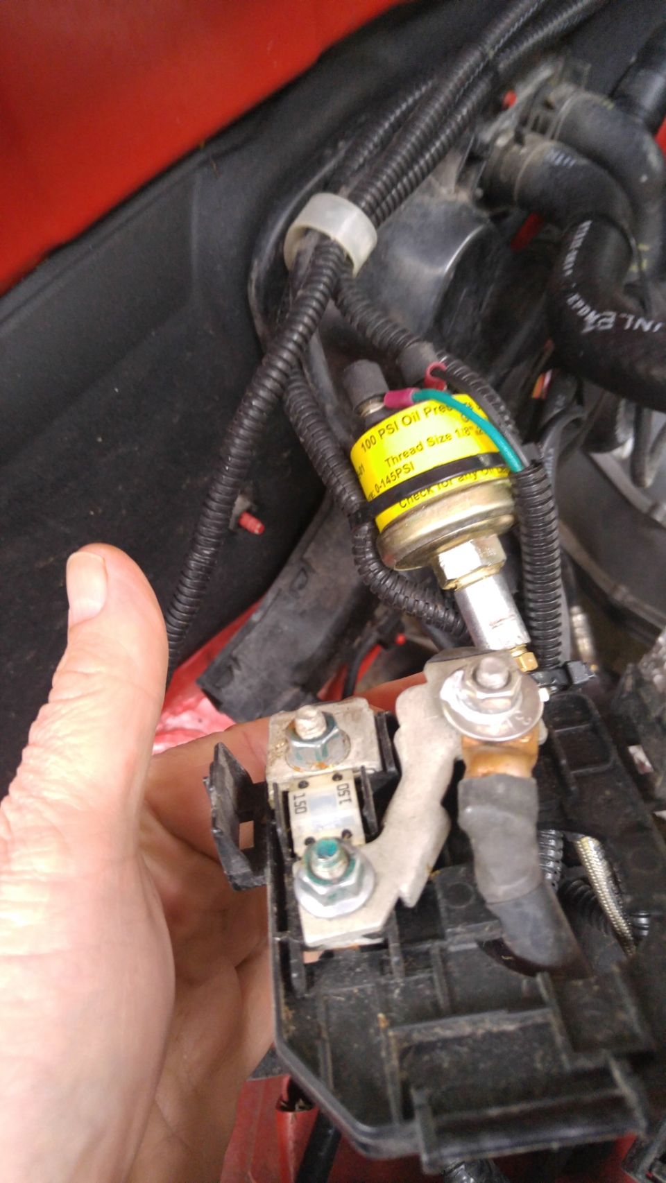 Hand holding fuse box with oil pressure sensor and wiring harnesses.