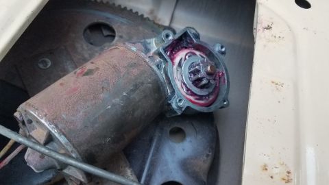 1985 Ford Bronco - A close-up view of a rusty power window motor with pink grease visible in the gears. The motor is mounted inside a vehicle door panel.