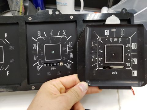 1978 Ford Bronco - A person's hand holds a black instrument cluster with white gauges, including a speedometer and odometer, against a larger black panel cont…