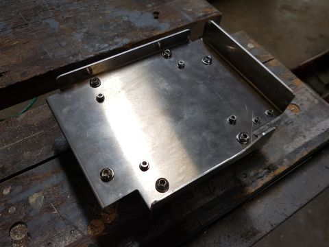 1978 Ford Bronco - A custom-fabricated stainless steel bracket with multiple nuts and bolts is shown resting on a weathered wooden surface. The bracket has a…