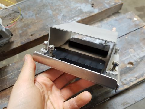 1978 Ford Bronco - A person's hand is holding a metal bracket with two black rectangular components attached to it. The bracket is secured by bolts and nuts t…