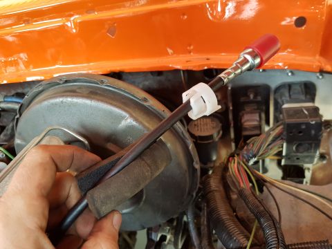 1978 Ford Bronco - A hand holds a black cable with a red cap on its end, secured by a white plastic clip, near a brake booster and engine wiring harness. The…