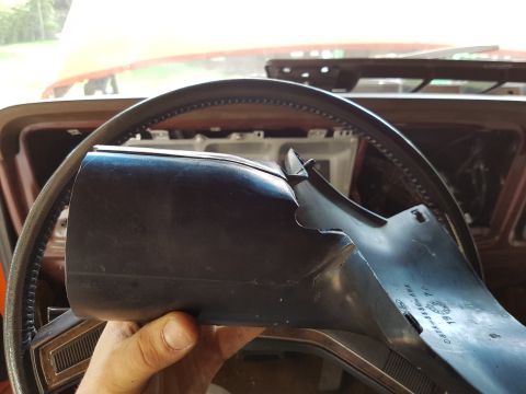 1978 Ford Bronco - A hand holds a cracked black plastic steering column cover in front of a steering wheel inside a vehicle. The cover has part numbers visibl…