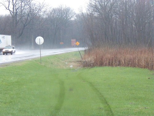 White sedan driving on a wet highway next to a semi-trailer truck, with trees and tall grass on the side