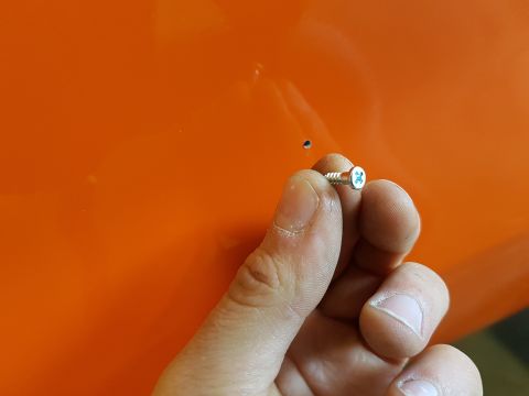 1978 Ford Bronco - A hand holds a screw next to a small hole on an orange surface.