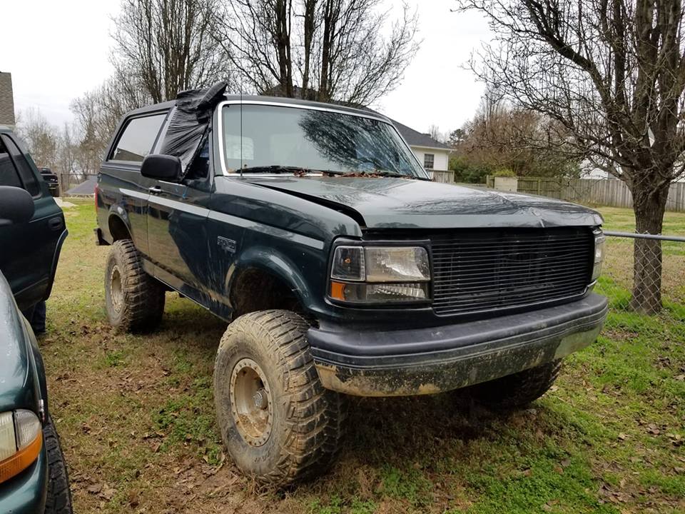 1993 Ford Bronco, dark green, lifted with large off-road tires. Black plastic covers driver's side window opening. Another vehicle is partially visible on the left