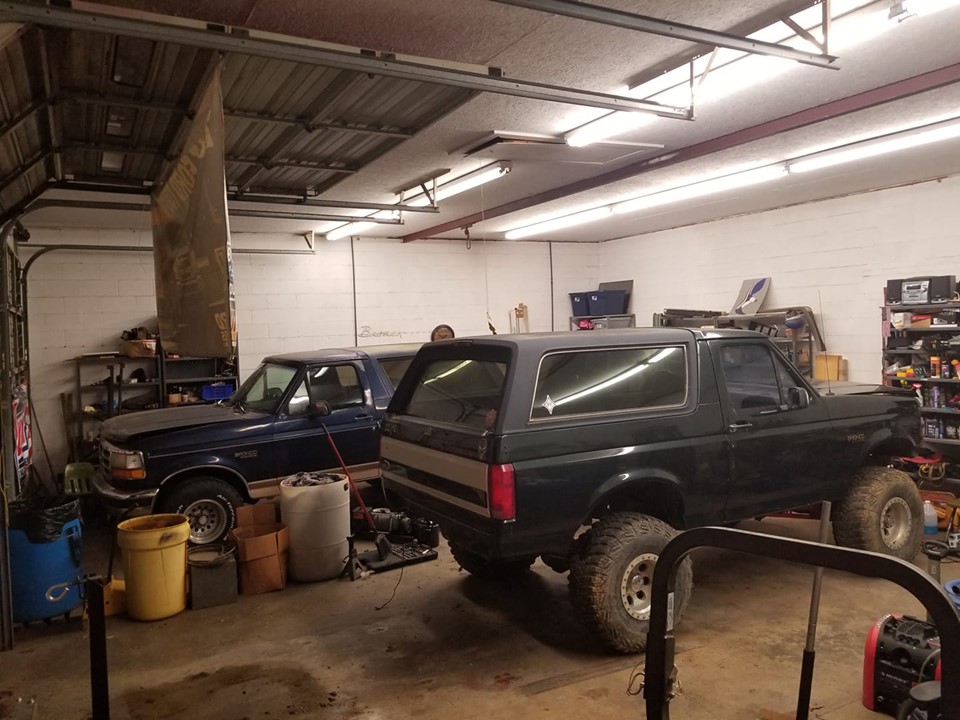 Dark green lifted Bronco and blue pickup truck parked in garage. Bronco has large off-road tires